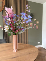 Un grand vase à rayures roses et blanches avec le bouquet de fleurs artificielles en soie Amethyst Romance se trouve sur une table ronde en bois dans une pièce moderne avec des murs verts, des boiseries et de petites œuvres d'art encadrées.