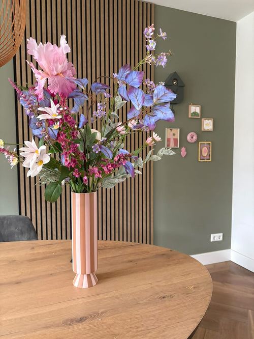 Un grand vase à rayures roses et blanches avec le bouquet de fleurs artificielles en soie Amethyst Romance se trouve sur une table ronde en bois dans une pièce moderne avec des murs verts, des boiseries et de petites œuvres d'art encadrées.