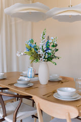 Une table à manger en bois recouverte d'assiettes, de bols, d'ustensiles de cuisine dorés et d'un vase blanc rempli du bouquet de fleurs artificielles en soie Cool Breeze. Un éclairage doux et des lampes suspendues blanches modernes au-dessus créent une atmosphère accueillante.