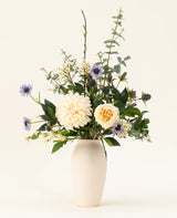 Un vase en céramique blanche contient le bouquet de fleurs artificielles en soie Petal Poetry, avec des dahlias et des roses de couleur crème, des fleurs violet clair, des chardons verts et toutes sortes de verdure sur un fond clair uni.