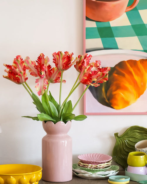 Sur une table avec de la vaisselle colorée se trouve un vase rose contenant la fleur artificielle tulipe de perruche française de 76 cm rouge/jaune, tandis qu'en arrière-plan on peut voir en partie une peinture encadrée d'un croissant et d'une tasse.