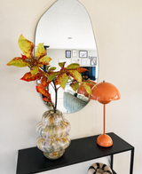Dans un miroir ondulé au-dessus d'une console noire se trouvent un vase en verre avec Kunsttak Chestnut 91 cm rouge/vert et une lampe champignon orange. Le miroir reflète un salon avec des œuvres d'art murales encadrées.