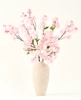 Le bouquet de fleurs artificielles en soie Fleur de cerisier rose 119 cm, disposé dans un vase en céramique beige aux délicates feuilles vertes, crée un accent intérieur serein sur un fond clair simple.