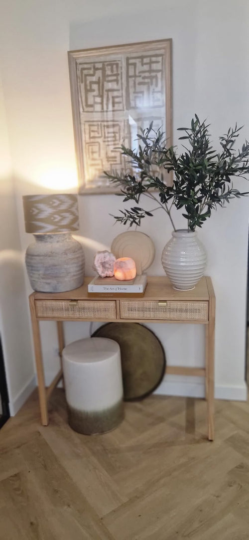 Une console en bois avec deux tiroirs contient une lampe, un grand vase blanc avec feuille d'olivier artificielle de 90 cm ignifuge, un plus petit vase, des livres et des pierres décoratives. Des accents méditerranéens et des œuvres d'art abstraites encadrées complètent le look ci-dessus. Au fond se trouvent un tabouret et un plateau rond.