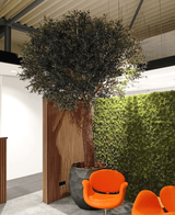 L'arbre artificiel Olivier 300 cm est situé à l'intérieur à côté d'un panneau en bois et d'un mur de mousse verte, avec deux chaises modernes orange devant lui sous un plafond industriel.