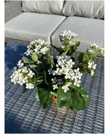 La fleur artificielle Kalanchoe UV de 31 cm de haut, avec des bouquets de petites fleurs blanches et des feuilles vertes, se dresse sur une table extérieure en osier avec un canapé gris avec des coussins en arrière-plan.