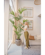 Dans un salon lumineux, une femme se tient à côté du palmier Kentia artificiel 220 cm, pendant qu'elle touche doucement les feuilles. L'intérieur tropical comprend un canapé lumineux avec des coussins, des œuvres d'art murales encadrées et de grandes fenêtres laissant entrer la lumière naturelle.