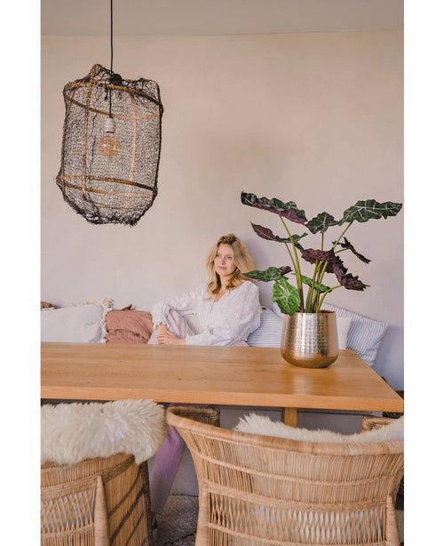 Une femme en blouse blanche est assise à une table à manger en bois avec des chaises en osier. La pièce confortable et doucement éclairée est dotée de la plante artificielle Alocasia UV 67 cm et d'une lampe suspendue texturée comme décor.