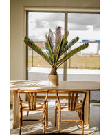 Une table à manger en bois avec deux chaises assorties se trouve près d'une grande fenêtre. Une plante artificielle Cycus Palm Deluxe de 95 cm se dresse sur la table et la lumière du soleil pénètre et projette ses ombres sur les meubles et le sol.