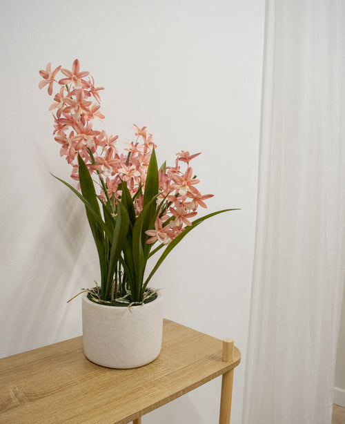 La plante artificielle Cymbidium rose de 58 cm, avec de hautes feuilles vertes et des fleurs roses ressemblant à des orchidées, se dresse sur une table en bois clair contre un mur blanc uni et un rideau blanc.