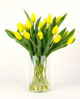 Le bouquet artificiel en soie Tulipes en vase Jaune se compose d'un bouquet de tulipes jaunes aux feuilles vertes dans un vase en verre transparent, sur un fond blanc uni.