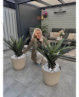 Une femme aux longs cheveux blonds s'agenouille et sourit entre deux plantes artificielles en pot Agava Pandora UV 85 cm sur une terrasse carrelée, avec des meubles d'extérieur et des fleurs sur le mur en arrière-plan.