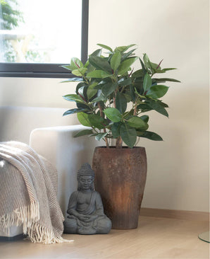 Une plante artificielle en caoutchouc de luxe de 90 cm se dresse sur un parquet en bois clair à côté d'une statue de Bouddha grise, tandis que la lumière du soleil tombe sur un canapé voisin drapé d'une couverture à franges beige et blanche.