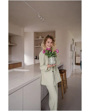 Une femme en costume de couleur claire sourit en tenant une fleur artificielle pétunia UV 25 cm rose foncé ; elle se tient dans une cuisine moderne aux tons neutres et à la décoration minimaliste.