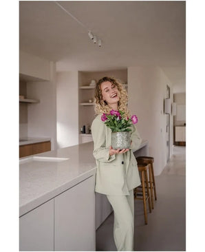 Une femme souriante aux cheveux bouclés, vêtue d'un costume de couleur claire, se tient dans une cuisine moderne et minimaliste tenant une fleur artificielle pétunia UV 25 cm rose foncé avec des fleurs rose vif.