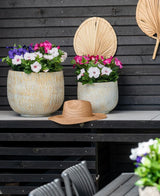 Deux grands pots en céramique avec une fleur artificielle Pétunia UV 25 cm rose foncé, avec des fleurs rose vif, se dressent sur une table contre un mur en bois sombre. Il y a un chapeau de paille sur la table et des feuilles de palmier séchées sont accrochées au mur au-dessus des fleurs.
