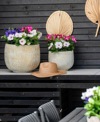 Deux grands pots en céramique avec une fleur artificielle Pétunia UV 25 cm rose foncé, avec des fleurs rose vif, se dressent sur une table contre un mur en bois sombre. Il y a un chapeau de paille sur la table et des feuilles de palmier séchées sont accrochées au mur au-dessus des fleurs.