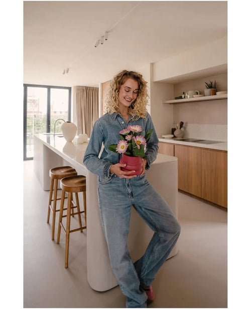 Une femme en jeans se tient debout dans une cuisine moderne, souriante et tenant dans ses mains la plante artificielle Gerbera UV rose de 28 cm – un petit pot de fleur rose avec des fleurs roses et blanches. Au fond, on aperçoit deux tabourets en bois et une grande fenêtre.