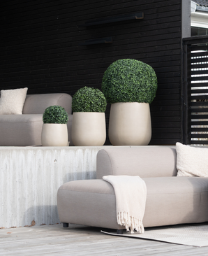 Un patio moderne comprend des coussins de siège beiges, une couverture crème et quatre plantes topiaires artificielles en buis romain de style romain UV D30 cm dans des pots de couleur claire contre un mur lambrissé en bois sombre.