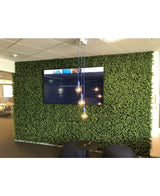 Sur un mur recouvert de panneaux d'Eucalyptus en haie artificielle de 50 x 50 cm (résistants aux UV), se trouvent une télévision, trois suspensions et une petite table ronde noire en dessous, avec un espace bureau en arrière-plan.