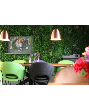 Un coin repas moderne avec des chaises vertes et pêche, une table décorée de vases de fleurs violettes, des suspensions en cuivre, un mur végétal luxuriant réalisé avec le panneau UV Kunsthaag Crush de 100 x 100 cm, et une photo encadrée d'un tigre en arrière-plan.