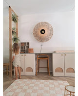 Une jeune fille en robe légère se tient à côté d'une étagère sur laquelle se trouvent la plante artificielle suspendue 'Frosted Fern on ball 76 cm' et d'autres plantes suspendues, dans une pièce moderne avec des placards beiges, un tabouret haut, une applique en osier et un tapis à carreaux au sol.