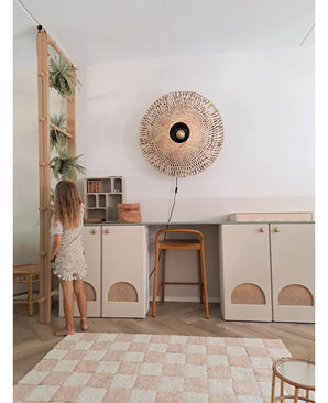 Une jeune fille en robe légère se tient à côté d'une étagère sur laquelle se trouvent la plante artificielle suspendue 'Frosted Fern on ball 76 cm' et d'autres plantes suspendues, dans une pièce moderne avec des placards beiges, un tabouret haut, une applique en osier et un tapis à carreaux au sol.