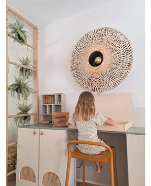 Une jeune enfant aux cheveux mi-longs est assise à un bureau dans une pièce lumineuse, le dos tourné au spectateur. Derrière elle se trouvent une applique murale tressée, quelques armoires et la plante artificielle suspendue « Fougère givrée sur boule 76 cm », nichée parmi les plantes sur une échelle en bois, à côté de petites étagères avec des objets bien rangés.