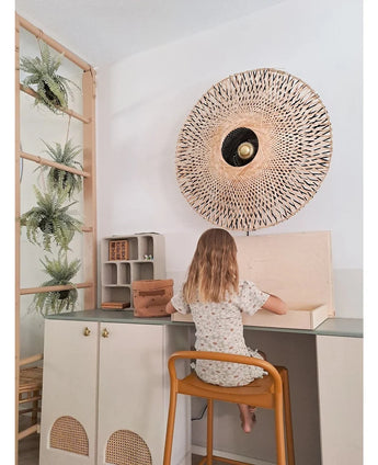 Une jeune enfant aux cheveux mi-longs est assise à un bureau dans une pièce lumineuse, le dos tourné au spectateur. Derrière elle se trouvent une applique murale tressée, quelques armoires et la plante artificielle suspendue « Fougère givrée sur boule 76 cm », nichée parmi les plantes sur une échelle en bois, à côté de petites étagères avec des objets bien rangés.