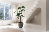 Chambre minimaliste et ensoleillée avec de grandes fenêtres, une plante artificielle Monstera de 210 cm, un tapis beige, un escalier blanc unique avec une niche voûtée en dessous. Des rideaux lumineux et une plante sur le rebord de la fenêtre complètent cet espace serein.