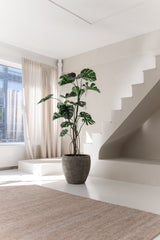 La plante artificielle Monstera 210 cm se dresse dans un pot sur un sol blanc à côté d'un escalier moderne dans une pièce lumineuse et minimaliste où la lumière naturelle pénètre à travers des rideaux beiges.