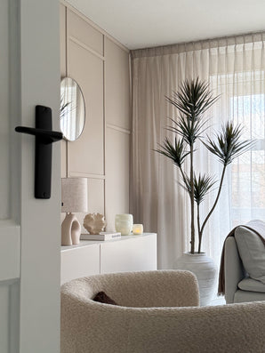 Un salon moderne et minimaliste avec un fauteuil texturé beige, une plante artificielle Dracaena Fragnans 180 cm, un buffet blanc décoré, un miroir rond et des rideaux transparents qui filtrent la douce lumière du jour à travers la fenêtre.
