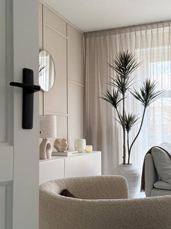 Un salon moderne et minimaliste avec un fauteuil texturé beige, une plante artificielle Dracaena Fragnans 180 cm, un buffet blanc décoré, un miroir rond et des rideaux transparents qui filtrent la douce lumière du jour à travers la fenêtre.