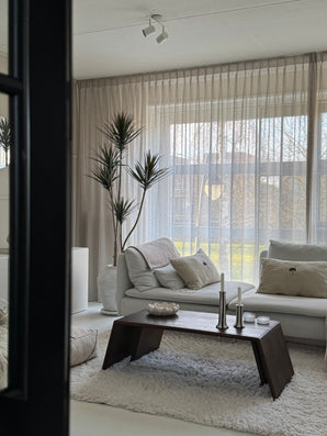 Un salon moderne avec de grandes fenêtres, des rideaux transparents, un canapé blanc avec coussins, une table basse en bois sombre avec bougies et décoration, la plante artificielle en pot Dracaena Fragnans 180 cm et un tapis blanc moelleux. Une douce lumière naturelle remplit la pièce.