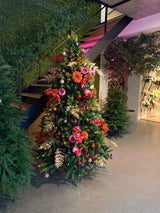 Le Sapin de Noël Artificiel "Weideman" PE/PVC - Smart LED est situé en intérieur près d'un escalier, décoré de fleurs roses et rouges, de feuilles dorées et d'ornements roses, entouré de verdure sur un sol en béton ciré pour une décoration de Noël stylée.