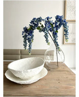 Une composition florale de Kunst Wisteria mesurant 115 cm se trouve dans un vase transparent sur une table en bois, à côté de deux bols blancs avec une structure, tandis que le mur derrière est décoré d'art abstrait.