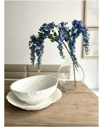 Une composition florale de Kunst Wisteria mesurant 115 cm se trouve dans un vase transparent sur une table en bois, à côté de deux bols blancs avec une structure, tandis que le mur derrière est décoré d'art abstrait.