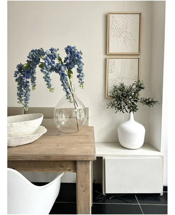 Sur une table en bois se trouve un vase transparent contenant la Glycine Artificielle de 115 cm, à côté d'un bol blanc. A proximité, un vase de verdure blanc repose sur un support blanc, sous deux œuvres d'art encadrées et texturées sur un mur clair.