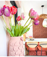 Un vase aux motifs de cœurs roses contient une fleur artificielle de tulipe française de 60 cm violette sur une table à manger avec des chaises aux dossiers tressés. En arrière-plan, un miroir rond et un mur à motifs marron contribuent à cet intérieur cosy et moderne.