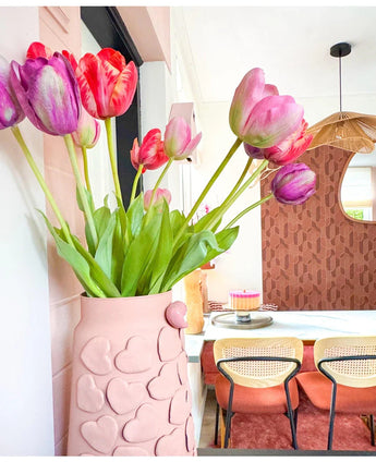 Un vase aux motifs de cœurs roses contient une fleur artificielle de tulipe française de 60 cm violette sur une table à manger avec des chaises aux dossiers tressés. En arrière-plan, un miroir rond et un mur à motifs marron contribuent à cet intérieur cosy et moderne.