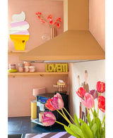 Une cuisine rose pastel avec une fleur artificielle de tulipe française de 60 cm violette au premier plan, une assiette LOVETTI jaune sur l'étagère, des tasses roses, une décoration murale représentant un cornet de glace et une fresque murale minimaliste représentant deux personnes au mur.