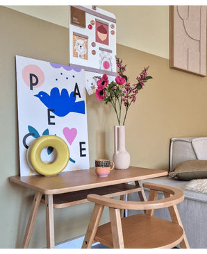 Un petit bureau en bois avec une chaise est décoré de fleur artificielle de cataire de 75 cm rose, d'une tasse, d'une sculpture en anneau jaune et de gravures d'art colorées sur le mur au-dessus. Un banc est partiellement visible à droite.