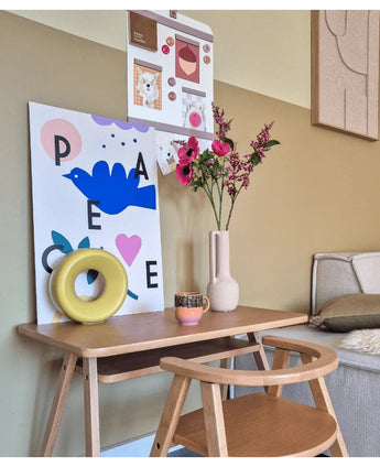 Un petit bureau en bois avec une chaise est décoré de fleur artificielle de cataire de 75 cm rose, d'une tasse, d'une sculpture en anneau jaune et de gravures d'art colorées sur le mur au-dessus. Un banc est partiellement visible à droite.