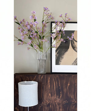 Dans un vase transparent, les fleurs du Kunsttak Daphne Spray 108 cm violet sont placées sur une surface en bois sombre à côté d'une œuvre d'art moderne et abstraite dans un cadre, avec un abat-jour blanc structuré en dessous.