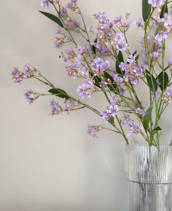 Branche artificielle raffinée Branche de Daphné de 108 cm de couleur violette, aux feuilles vertes, disposée dans un vase en verre transparent nervuré et posée sur un fond uni de couleur claire.