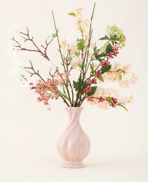 Un vase en céramique rose contient le bouquet de fleurs artificielles en soie Springtime Poem, avec des fleurs blanches, roses et vertes avec des feuilles vertes et des tiges brunes, sur un fond clair.