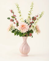 Een roze vaas van keramiek bevat het Zijden kunstbloemen boeket Morning Whisper, met lichtroze dahlia's, witte leeuwenbekjes, groene bladeren en trosjes kleine rode besjes, tegen een effen gebroken witte achtergrond.