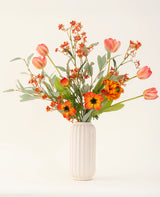 Le bouquet de fleurs artificielles en soie Orange Glory, avec des tulipes orange, des coquelicots et des feuilles vertes, est magnifiquement disposé dans un vase blanc texturé sur un fond clair uni.