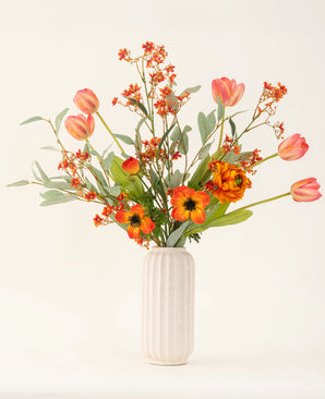 Le bouquet de fleurs artificielles en soie Orange Glory, avec des tulipes orange, des coquelicots et des feuilles vertes, est magnifiquement disposé dans un vase blanc texturé sur un fond clair uni.