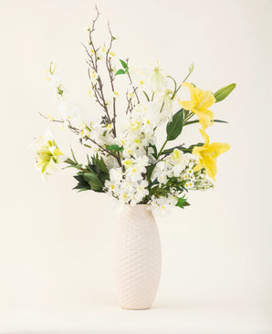 Un vase à structure blanche montre l'élégance du bouquet de fleurs artificielles en soie Sunkissed Ivory, avec des lys jaunes, des fleurs blanches et des feuilles vertes sur un fond uni clair.